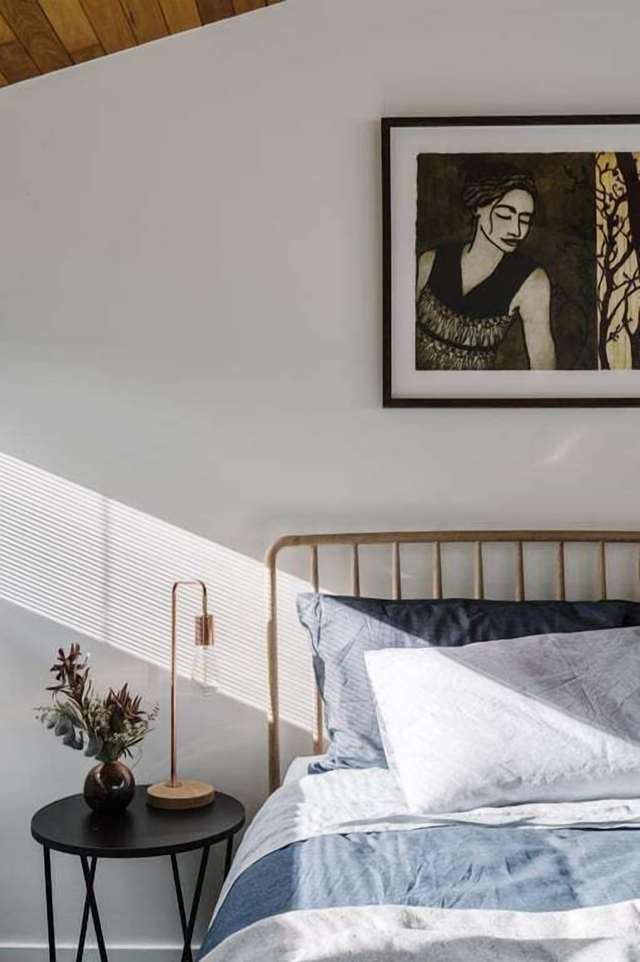 Un dormitorio con cama, una obra de arte enmarcada en la pared, una mesita redonda con una planta y una ventana con persianas.