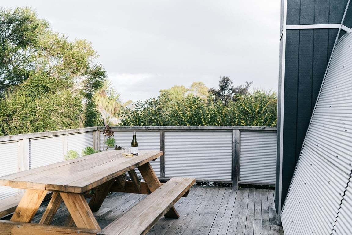 Une terrasse en bois avec une table de pique-nique et une bouteille de vin, entourée d’une clôture en métal et de verdure.