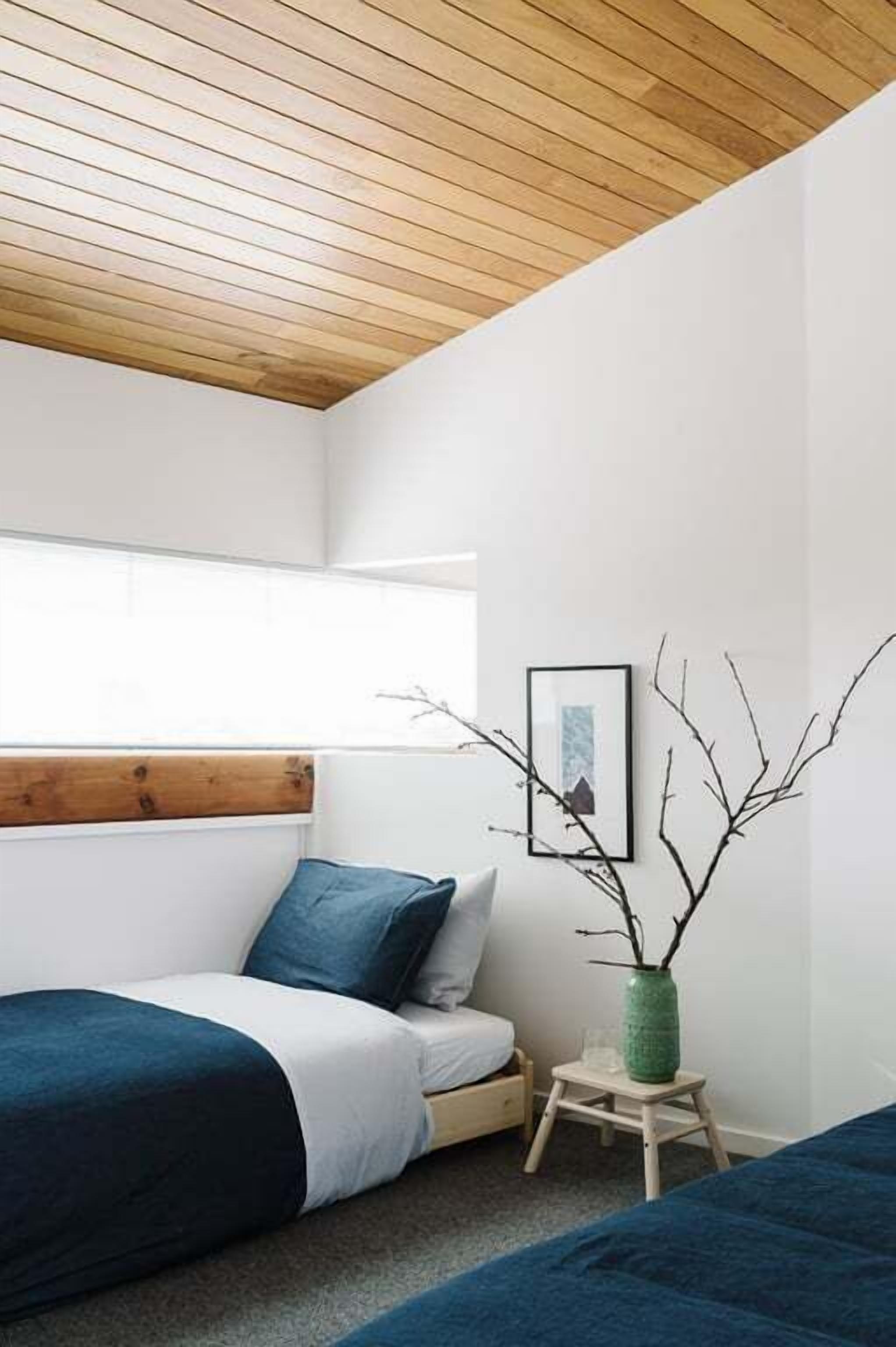 Quarto com teto de madeira, cama com roupa de cama azul, um banquinho de madeira e um vaso verde com galhos.