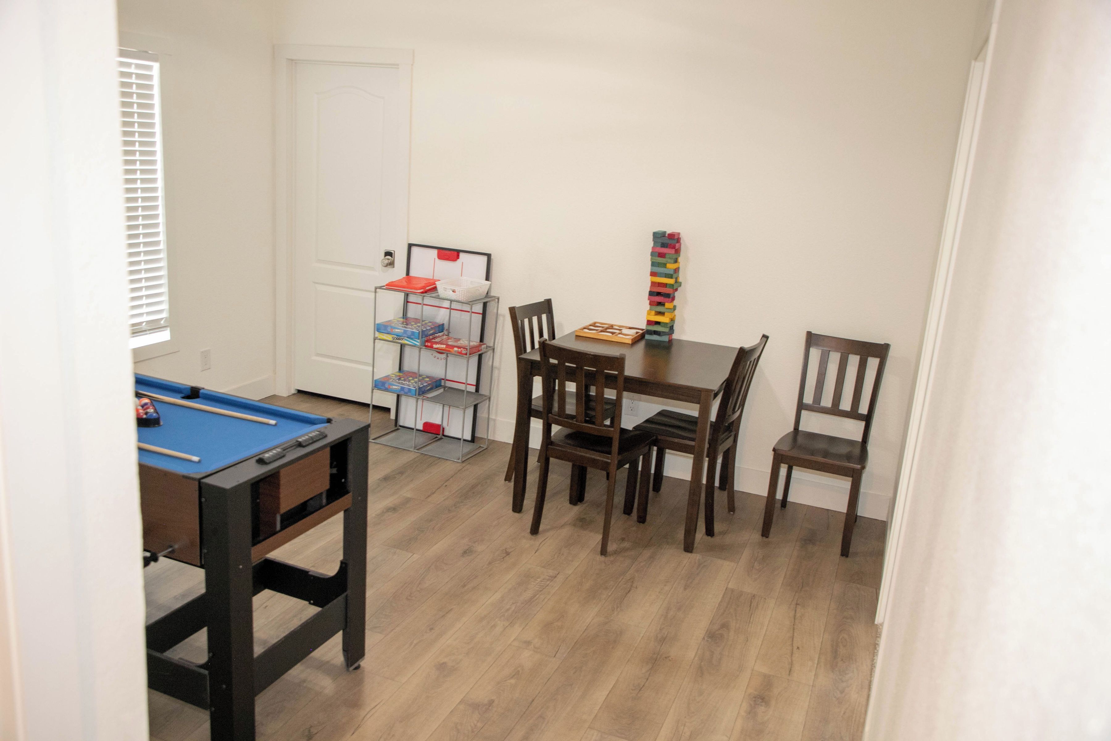 Una habitación con mesa de billar, mesa de comedor con sillas, estantería y ventana con persianas.