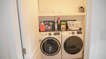 A laundry room with two front-loading washing machines, a shelf with cleaning supplies, and a basket.