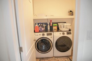 A laundry room with two front-loading washing machines, a shelf with cleaning supplies, and a basket. - Enjoyable 4-bedroom house with AC in charming Taylor.  (Taylor)