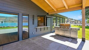A modern outdoor kitchen with a built-in grill, large windows, and a view of a lake.