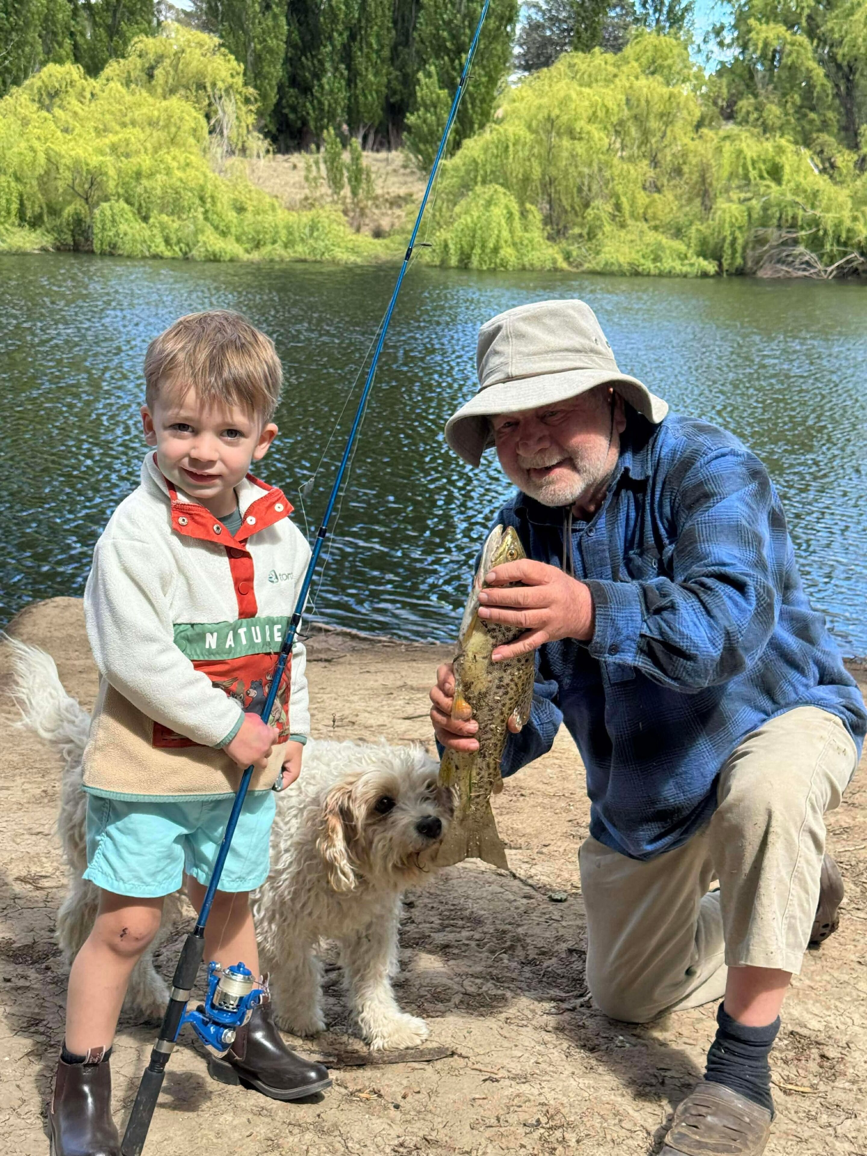 Eine Person hält einen Fisch in der Nähe von Gewässer, ein Hund ist in der Nähe.