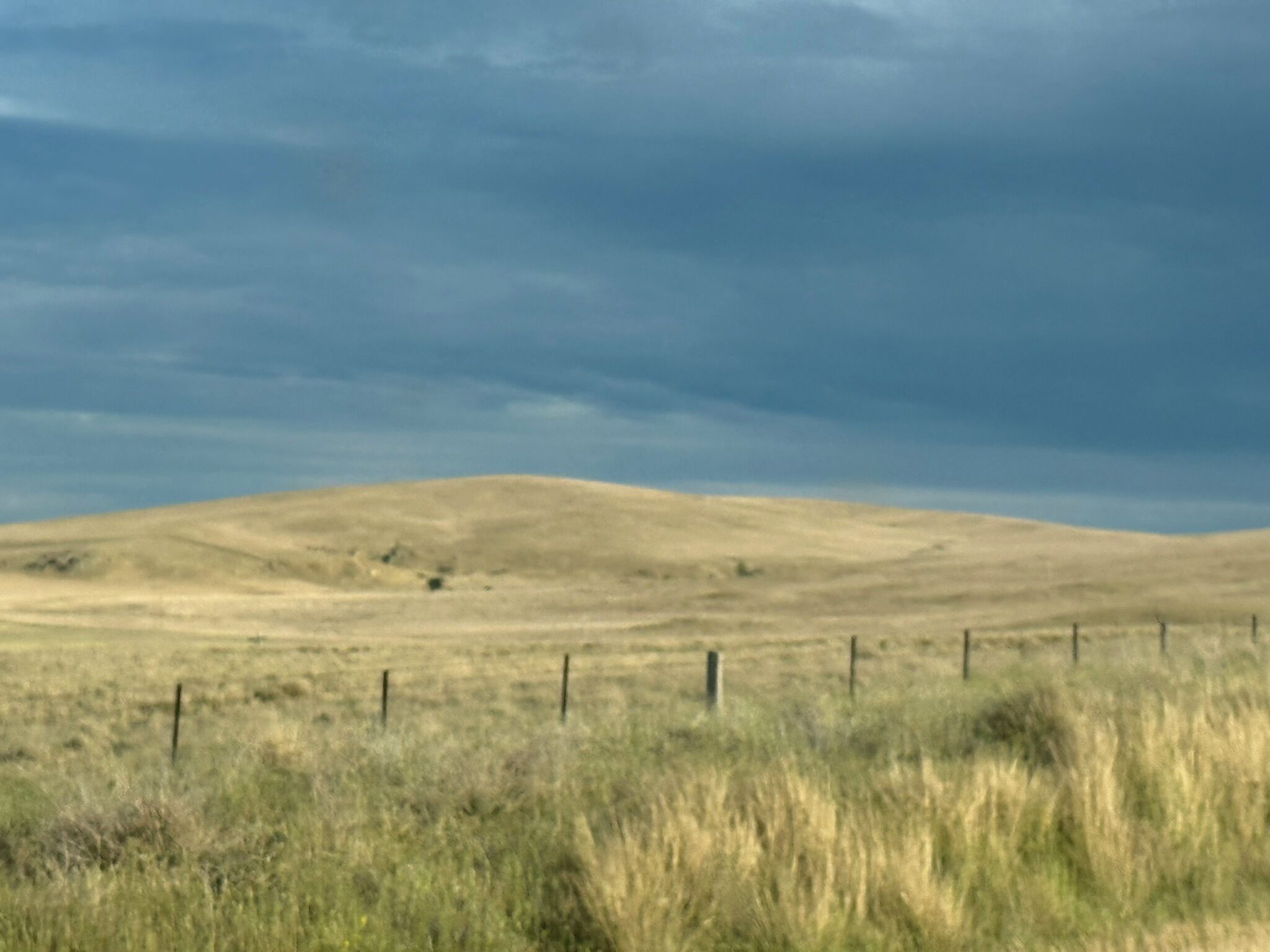 A flat, open landscape with a fence in the foreground and a hill in the background under a cloudy sky.