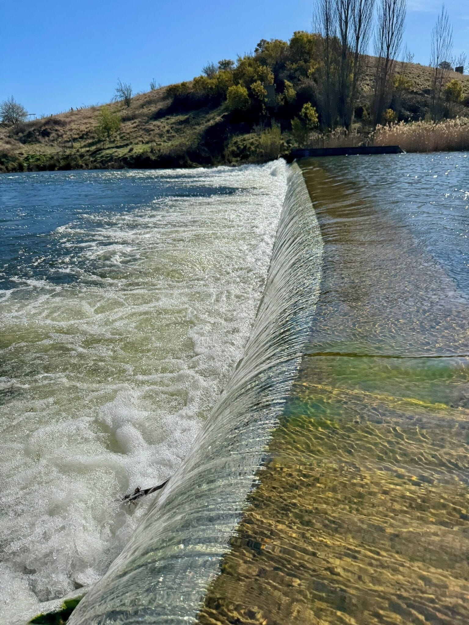 A river with a weir, clear water, and a hilly landscape with sparse vegetation.