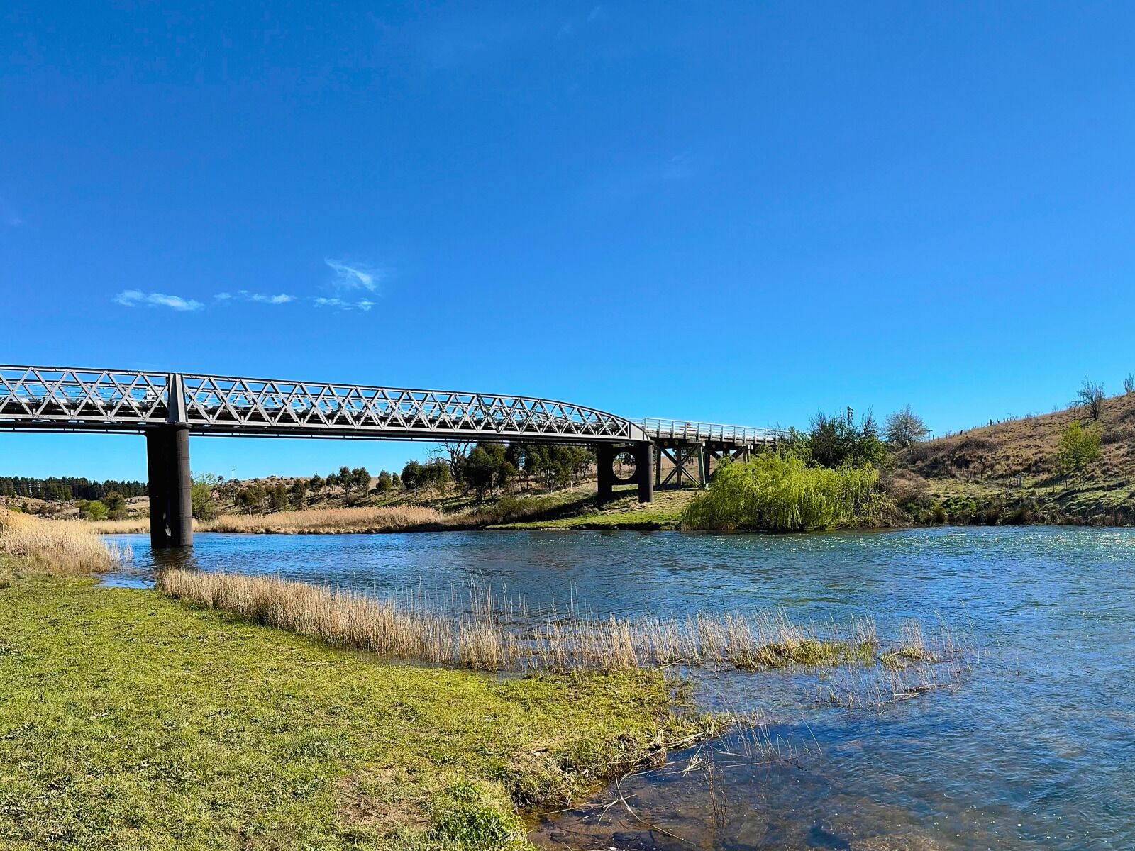 Un puente de metal sobre un río, con orillas cubiertas de hierba y árboles al fondo.