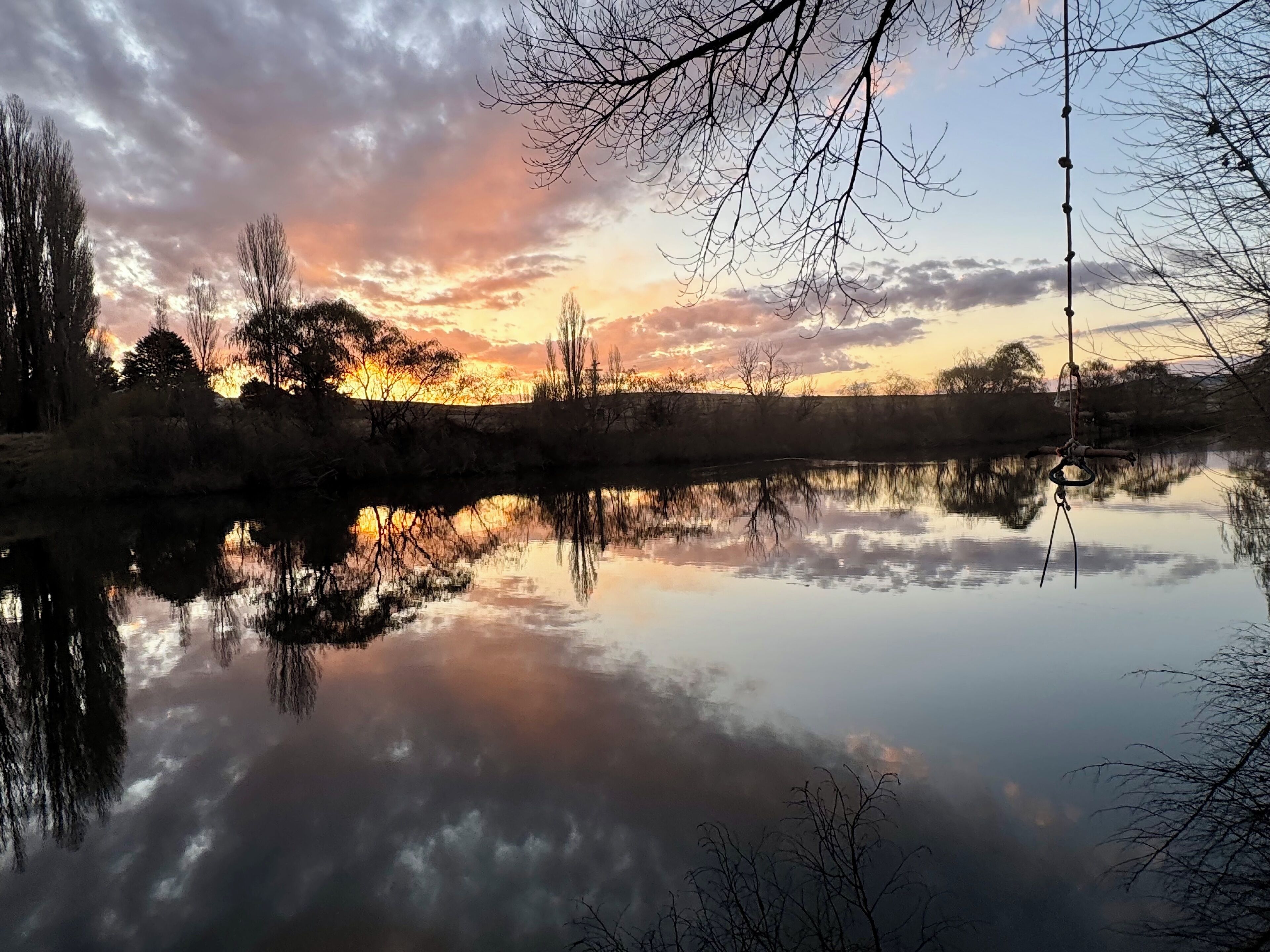 Un llac tranquil que reflecteix el cel, amb arbres i una vara de pescar en primer pla.