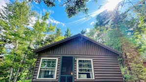 Une cabane avec une porte d’entrée, deux fenêtres et une terrasse en bois.