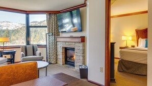 Quarto de hotel com lareira de pedra, televisão na parede, cama e vista para as montanhas através de grandes janelas.