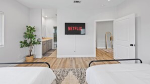 Quarto de hotel moderno com uma cama, uma mesinha de cabeceira, um vaso de planta e uma TV na parede exibindo 'NETFLIX'.
