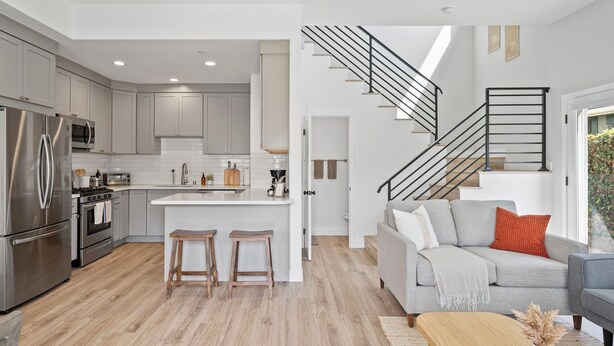 Cozinha moderna com eletrodomésticos de aço inoxidável, uma ilha central com banquetas e uma escadaria que dá acesso a uma sala de estar com sofá cinza e mesa de centro.
