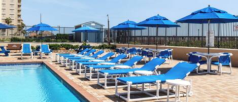 A poolside area with lounge chairs and umbrellas, a swimming pool, and a building in the background.