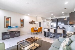 A modern living room with a flat-screen TV, dining table, and chairs, a kitchen with bar stools, and a staircase leading to an upper floor. - 1521 Dream (Clermont)