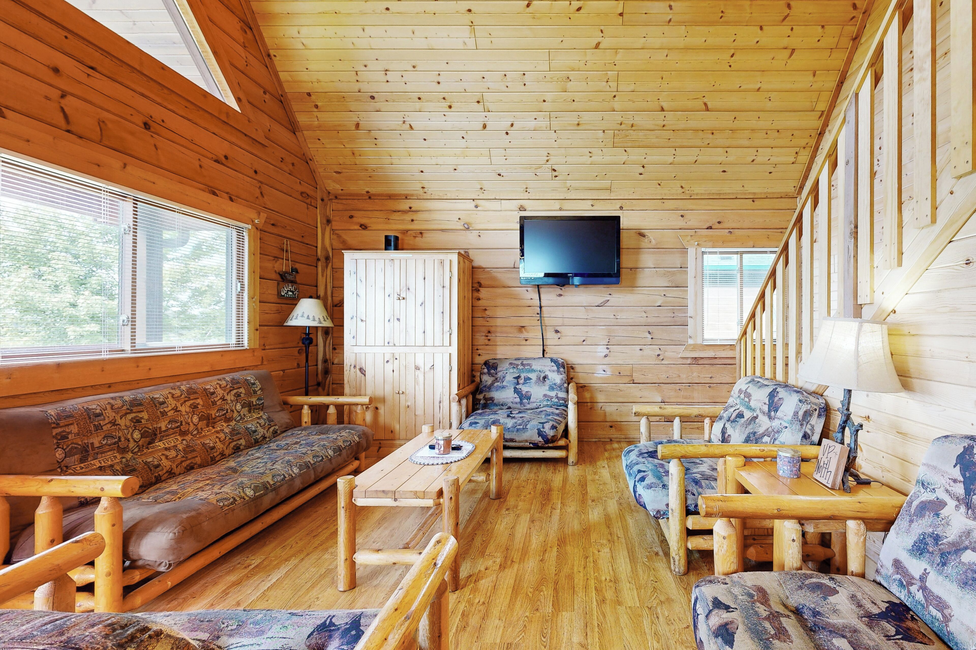 A wooden cabin interior with a sofa, armchairs, a table, and a television mounted on the wall.