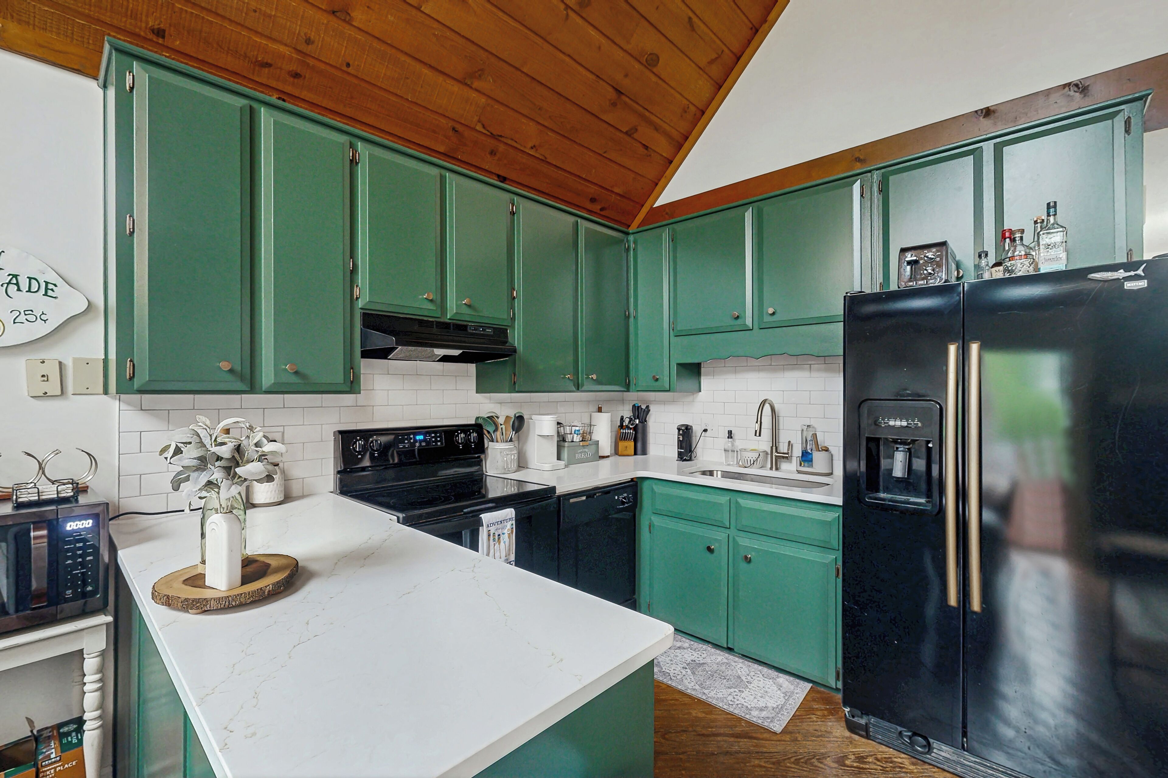 Une cuisine avec des meubles de couleur verte, un réfrigérateur noir et un plan de travail blanc.