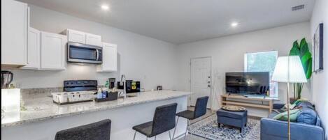 Cocina moderna con armarios blancos, microondas y estufa. Hay un comedor con un taburete de bar y un sofá. Una planta grande se encuentra a la derecha.