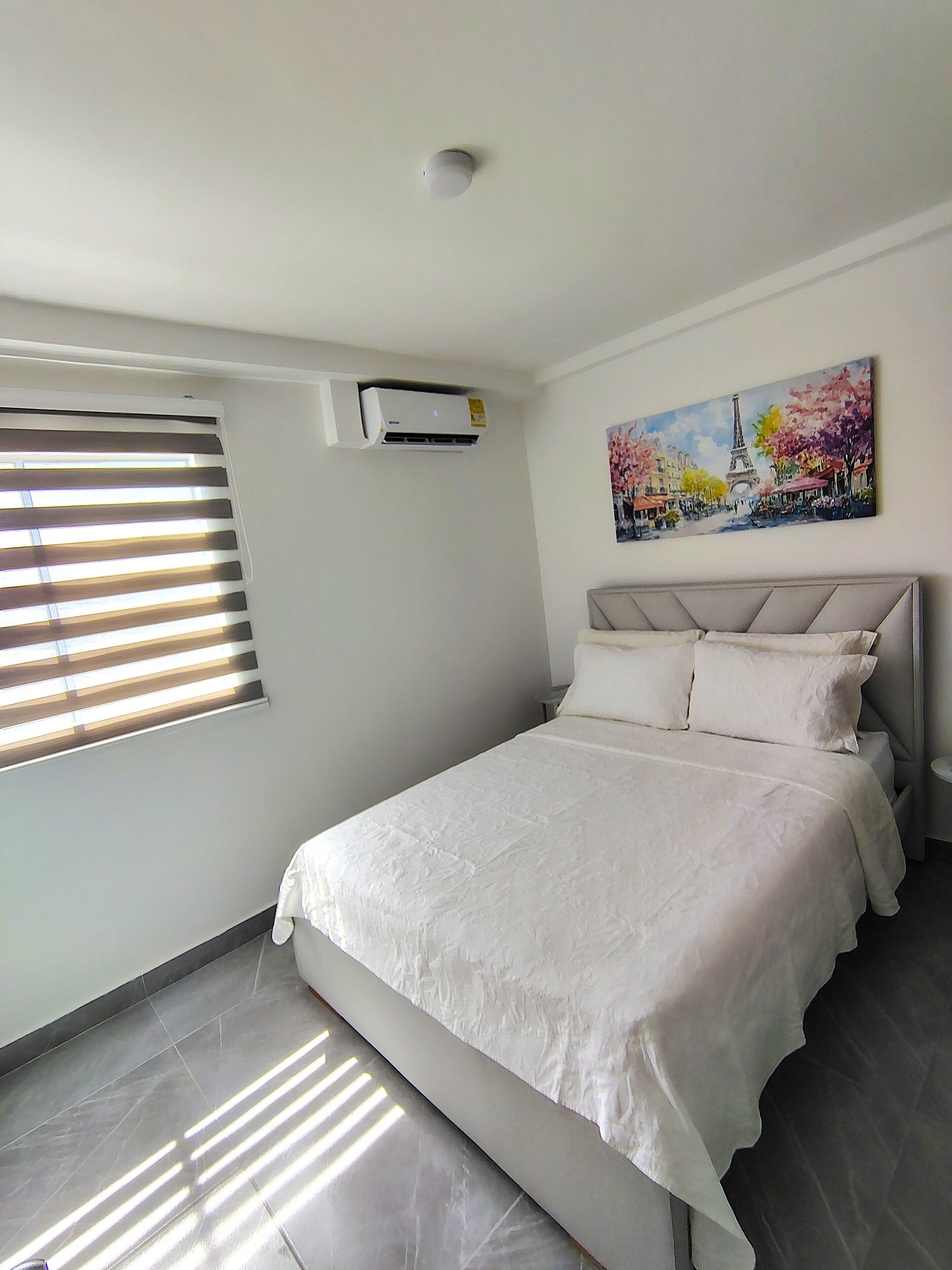 A bedroom with a bed, a window with blinds, an air conditioning unit, and a painting of a Parisian street scene.
