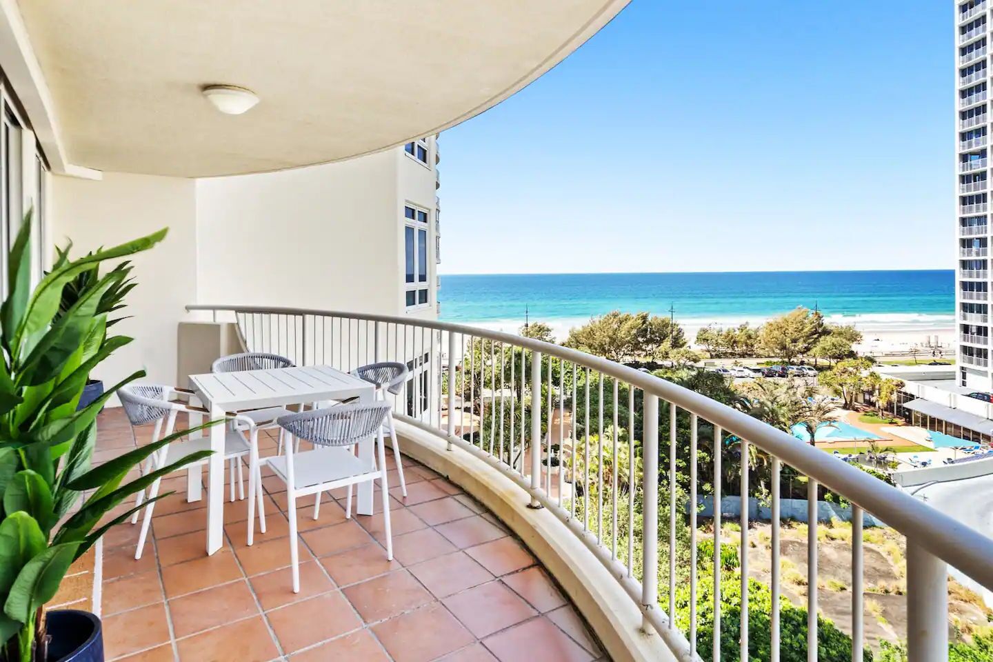 Een balkon met een witte tafel en stoelen, uitkijkend op een strand met een helder uitzicht op de oceaan.