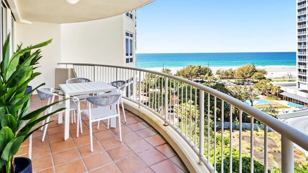 Un balcon avec une table et des chaises blanches, donnant sur une plage et offrant une vue dégagée sur l’océan.