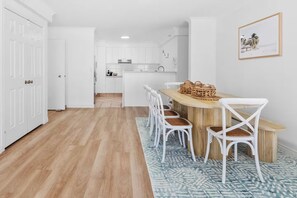 A modern kitchen with white cabinets, a wooden dining table with white chairs, and a blue patterned rug. - Large 3 Bedroom Ocean Views (Surfers Paradise)