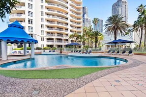 A swimming pool with lounge chairs and umbrellas, surrounded by a landscaped area with palm trees. - Large 3 Bedroom Ocean Views (Surfers Paradise)