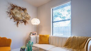 Un salon moderne avec un canapé blanc, une table basse en verre, un élément décoratif mural et une fenêtre avec des stores.