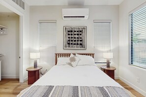 Une chambre avec un grand lit, deux tables de chevet avec des lampes, un climatiseur fixé au mur et une fenêtre avec des stores.