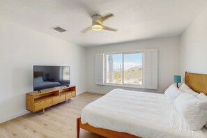 Une chambre à coucher avec un lit, un téléviseur posé sur un meuble en bois, un ventilateur de plafond, une fenêtre avec des volets et une table de chevet avec une lampe.