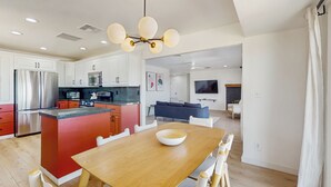 Eine moderne Küche mit einer zentralen Kochinsel, ein Essbereich mit Holztisch und ein Wohnzimmer mit Sofa und Fernseher.