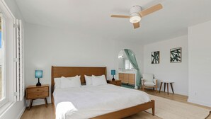 Ein Schlafzimmer mit einem hölzernen Bett, einem Deckenventilator, einem Nachttisch mit Lampe, einem Sessel und Bildern an der Wand.