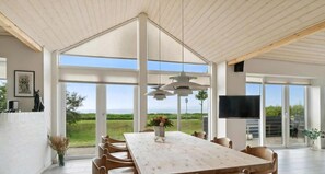 A spacious dining area with a large table and chairs, a TV mounted on the wall, and large windows offering a view of the outdoors. - Bright and lovely seaside holiday home (Ebeltoft)