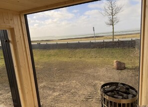 A view from a wooden-framed window overlooking a beach with a fence, a tree, and a fire pit. - Bright and lovely seaside holiday home (Ebeltoft)