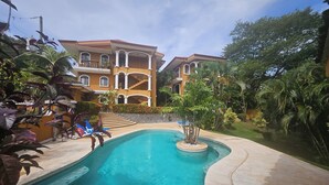 A pool area with a clear blue pool, surrounded by lush greenery and a large building with multiple balconies. - Main floor, Family unit, 3 bed, fresh, clean, inside Tamarindo w incredible pool (Tamarindo)