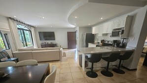 A modern kitchen with white cabinets, a central island with bar stools, a dining area with a table and chairs, and a living room with a sofa and a TV. - Main floor, Family unit, 3 bed, fresh, clean, inside Tamarindo w incredible pool (Tamarindo)