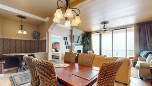Ein Essbereich mit Holztisch und gewobenen Stühlen, eine Küche im Hintergrund und ein Balkon mit Vorhängen.