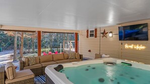A hot tub with a view of trees, a sofa, and a TV mounted on the wall. - KnoxHaven | 5 Min UT•Hot Tub•Firepit•Theater•Games (Knoxville)
