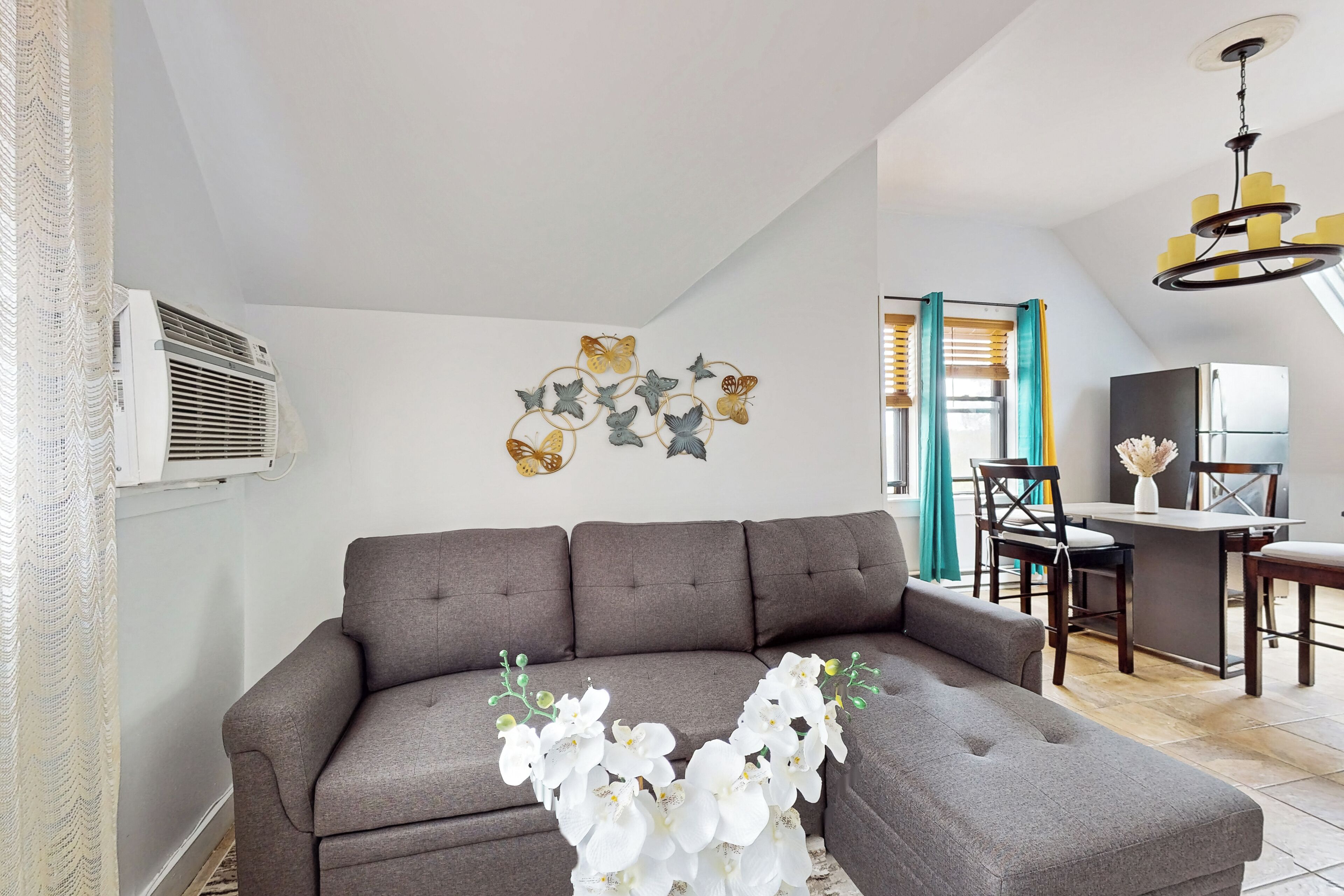 A living room with a grey sectional sofa, a wall-mounted air conditioner, and a decorative butterfly wall art piece.