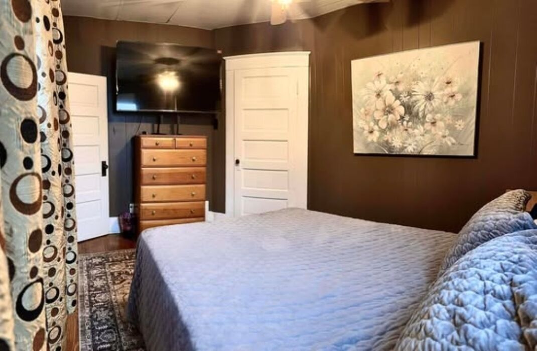 Une chambre à coucher avec un lit, une commode, un téléviseur fixé au mur et un tableau.