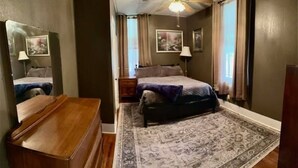 Ein Schlafzimmer mit einem Bett, einer Holzkommode, einem Deckenventilator, einem Teppich und einem Fenster mit Vorhängen.