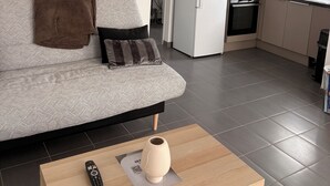 A modern living room with a grey sofa, a remote control, and a vase on a wooden coffee table. A kitchen with a fridge, microwave, and sink is visible in the background. - House near Bordeaux, 3 minutes from the ARENA (Bouliac)