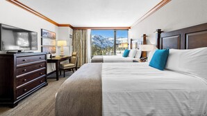 A hotel room with two beds, a desk, a TV, and a view of mountains.