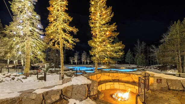 A stone fire pit surrounded by snow, with illuminated trees and a pool in the background.
