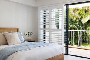 A bedroom with a bed, a nightstand, a small table with a plant, and a balcony with a view of palm trees.