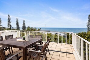 An outdoor patio with a wooden table and chairs, a white railing, and a view of the sea.