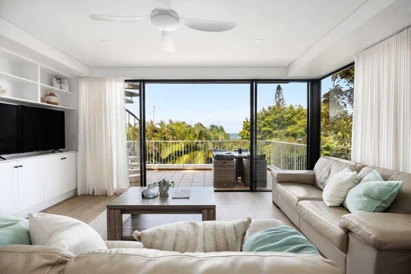 Ein modernes Wohnzimmer mit Fernseher, einer Couch, einem Couchtisch und einem Balkon mit Blick auf Grünflächen und einen klaren Himmel.