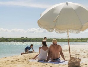 A beach scene with a large umbrella, people relaxing on the sand, and a body of water in the background. - Robert Street River Retreat 30 Secs to River (Noosaville)