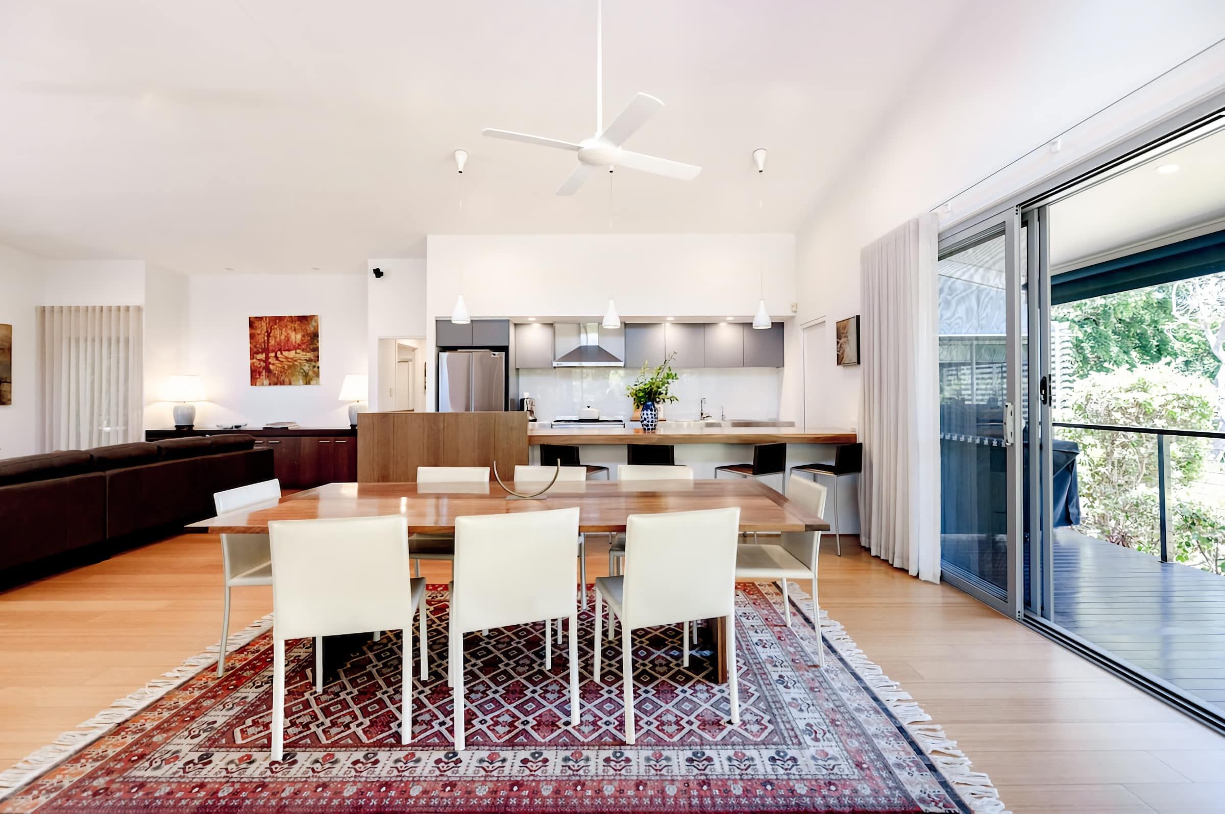 Una cocina moderna con una isla central, una zona de comedor con mesa de madera y sillas blancas, y un amplio ventanal corredero que da a un balcón.