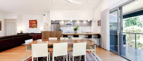 Een moderne keuken met een centraal eiland, een eethoek met een houten tafel en witte stoelen, en een grote schuifpui die leidt naar een balkon.