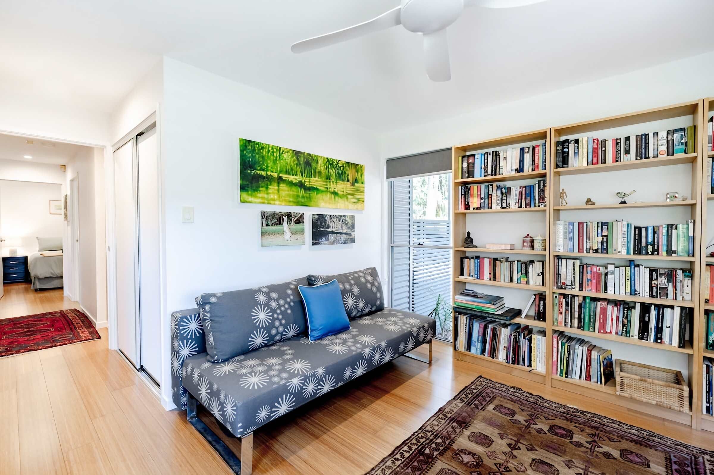 A modern living room with a grey sofa, a bookshelf filled with books, and a large window with blinds.